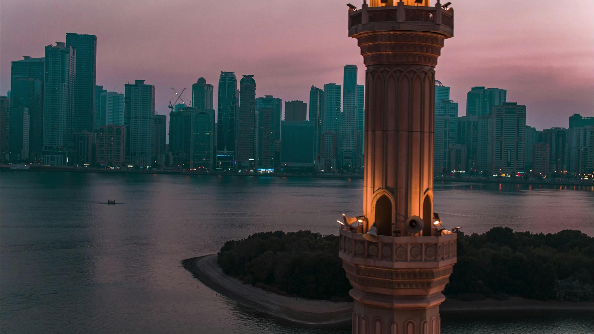 Sharjah's skyline across the lagoon at dusk, beside a lit minaret