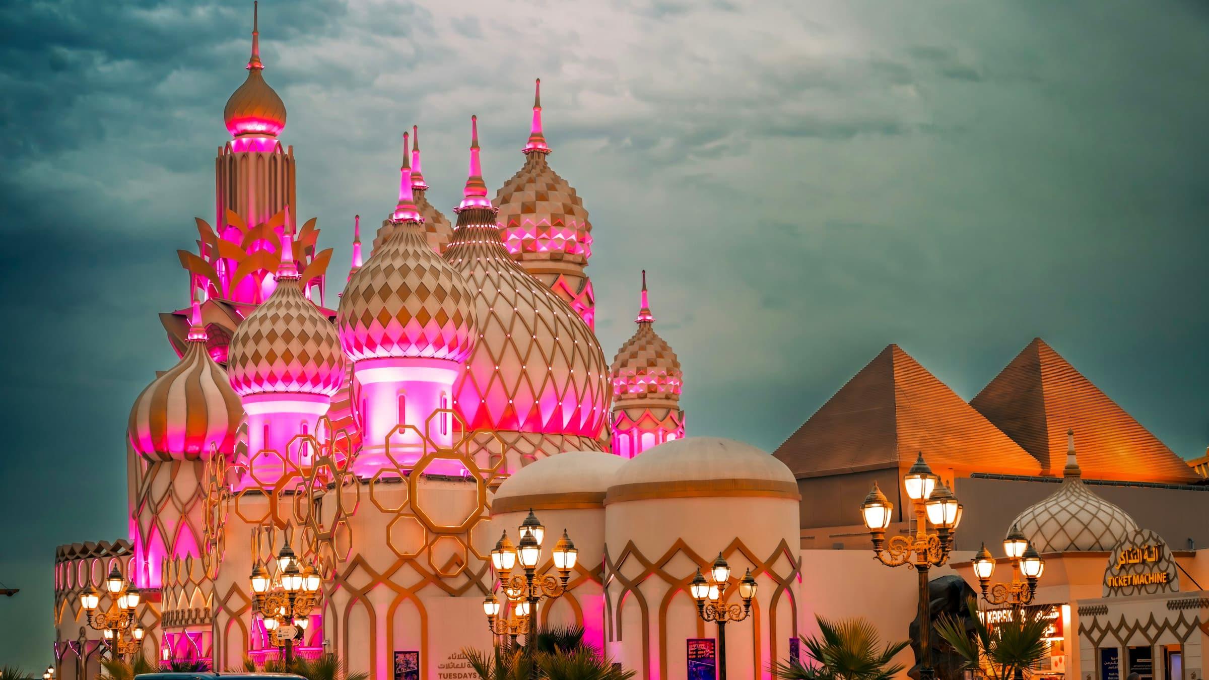 Pink-lit domes at the entrance to Global Village Dubai at dusk, with the park's pyramids behind