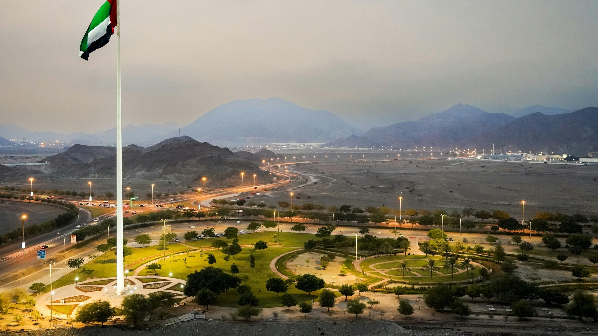 The UAE flag above Fujairah city, with the Hajar Mountains behind at dusk