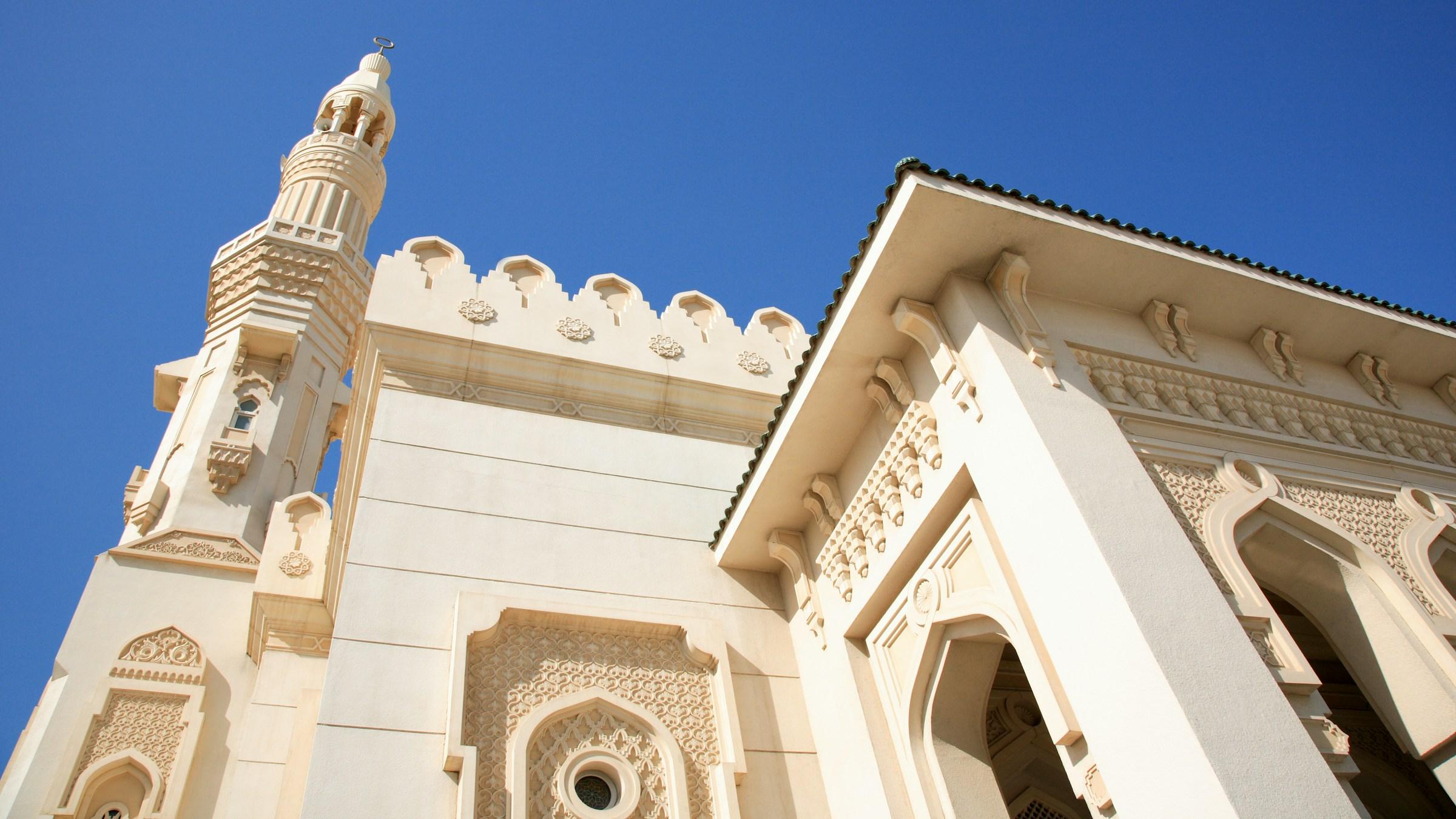A white mosque in Sharjah with a carved minaret and arched entrances against a blue sky