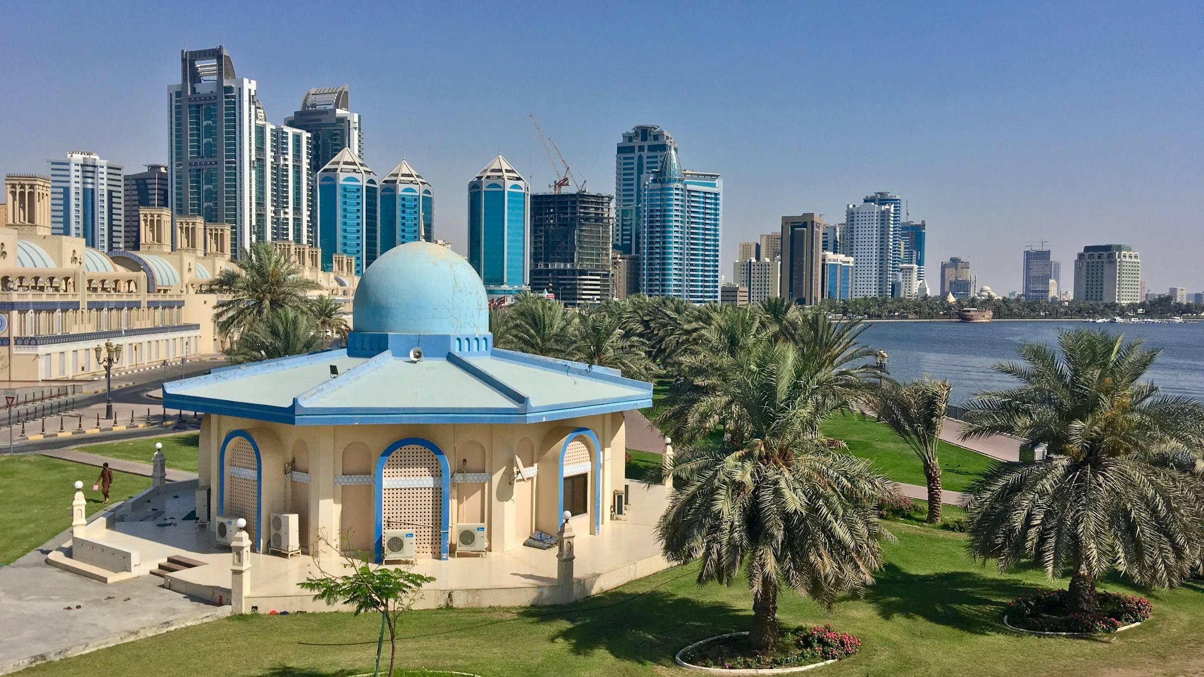 A blue-domed pavilion among palms in Sharjah, with the city's towers behind