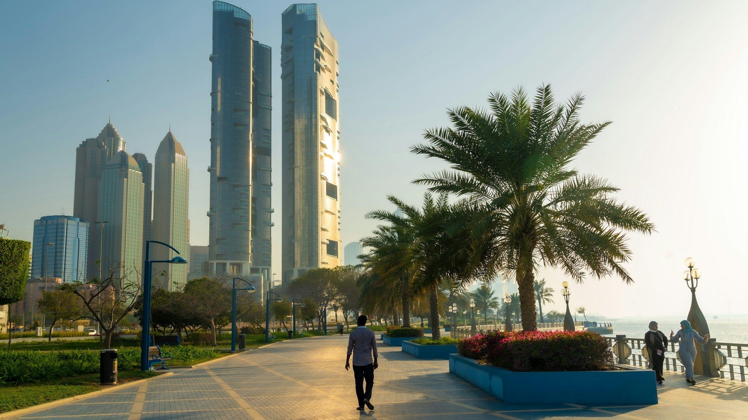 A walker on the Abu Dhabi Corniche promenade, with palms and city towers behind