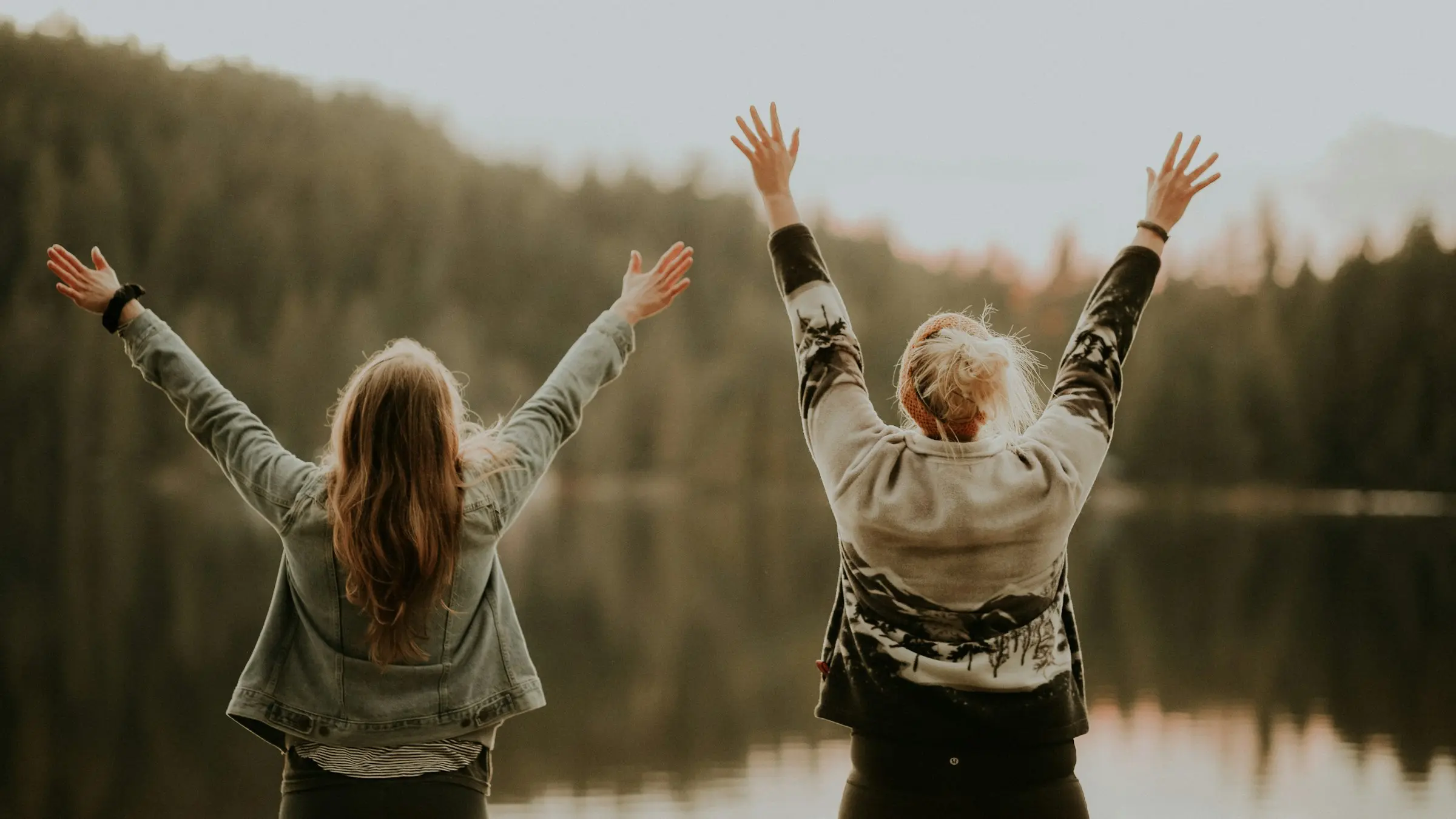Two women with their arms raised beside a lake at sunset