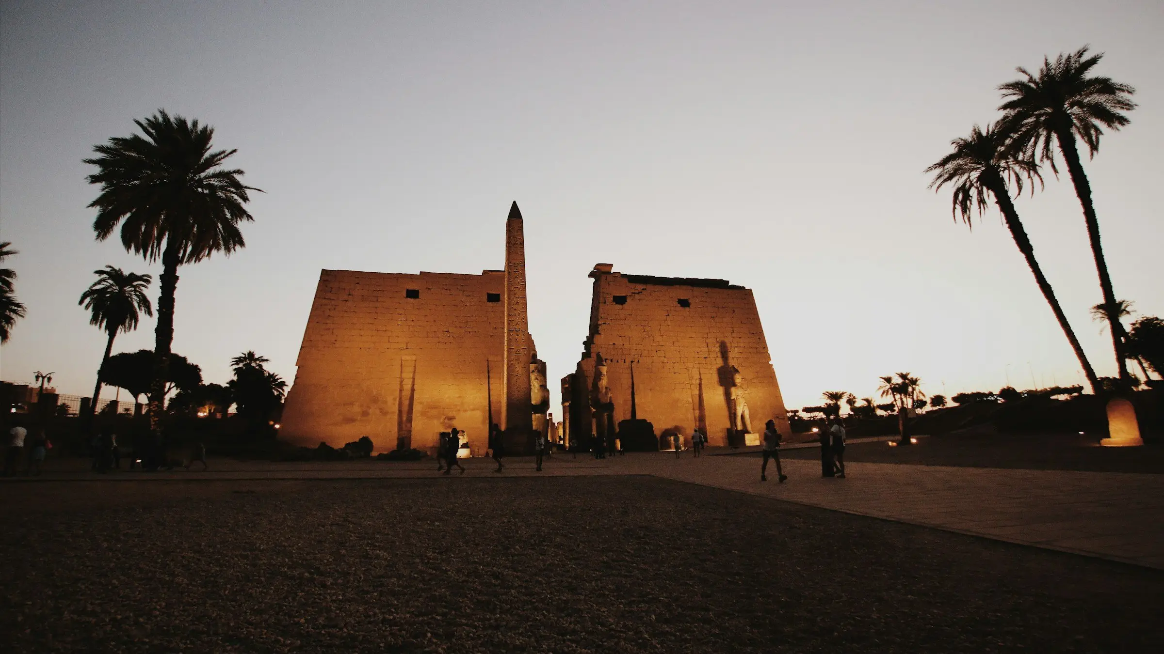 The pylon and obelisk of Luxor Temple lit at dusk, with palms on either side