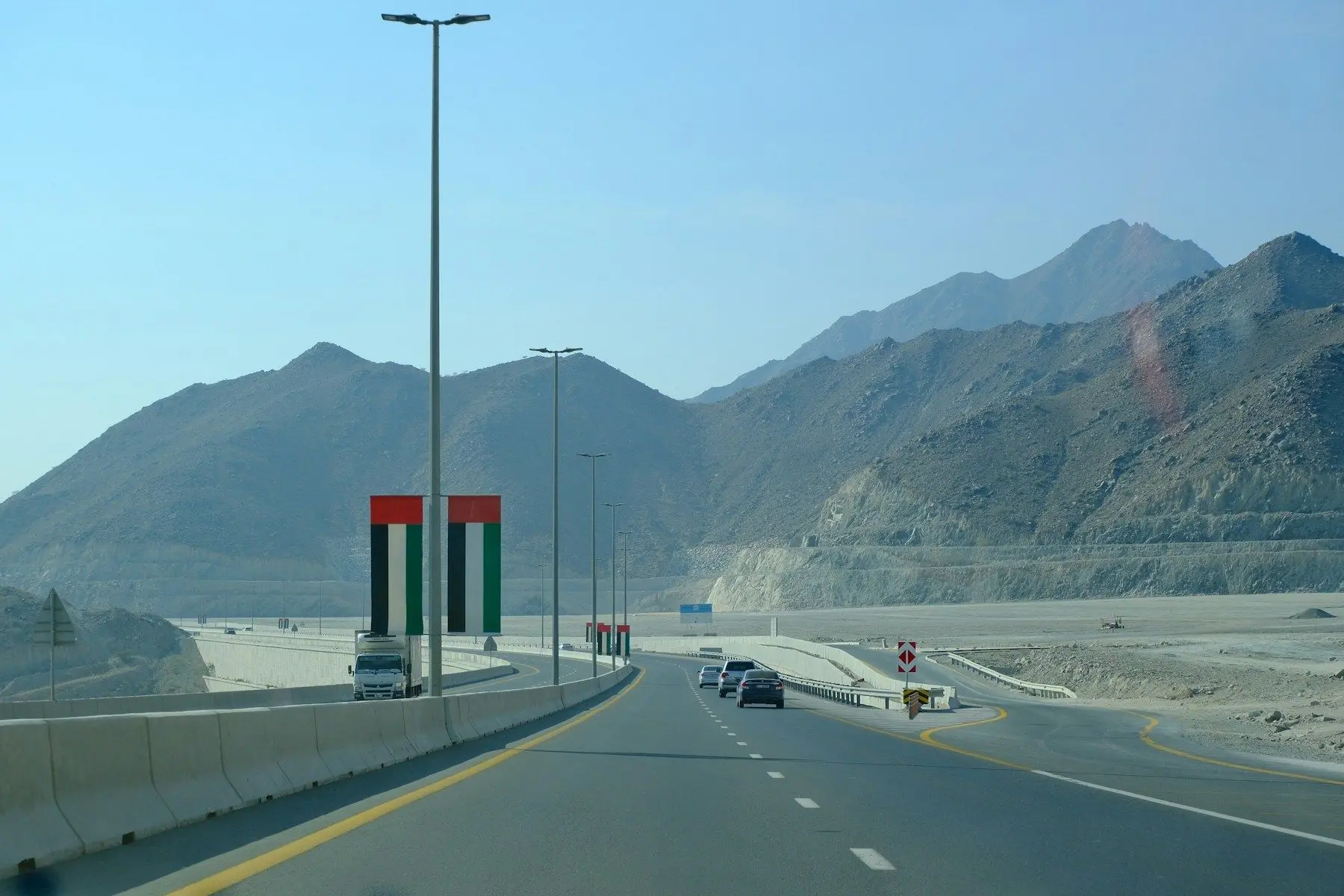 A highway through the Hajar Mountains past a monument in the colours of the UAE flag